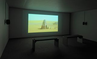 White darkened room, grey floor, two stool seats centre, projection screen on the wall of a desert, rock mound with a doorway and a line of four people in desert clothing walking out into the surroundings, blue hazy sky