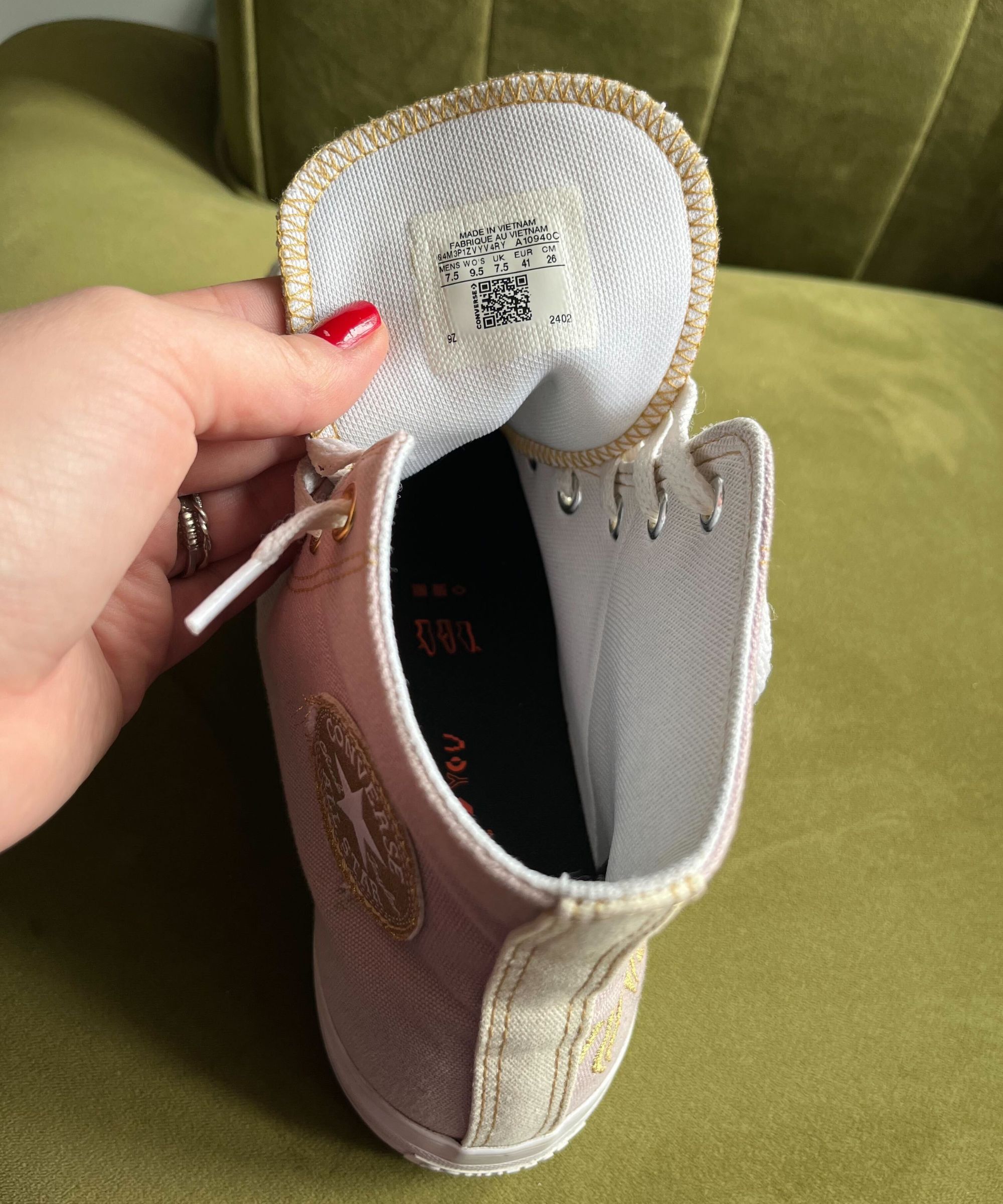 A light lilac high-top pair of lace up converse shoes on a green velvet sofa. There is a hand in view, holding back the tongue of the shoes, to reveal a QR code for care instructions