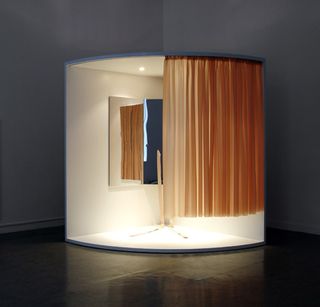Strap-in Mirror Corner, by Anna + Peter, 2010. A quarter circle shaped cubicle with mirrors inside it, glowing lights inside of it and curtains running across the front of it.