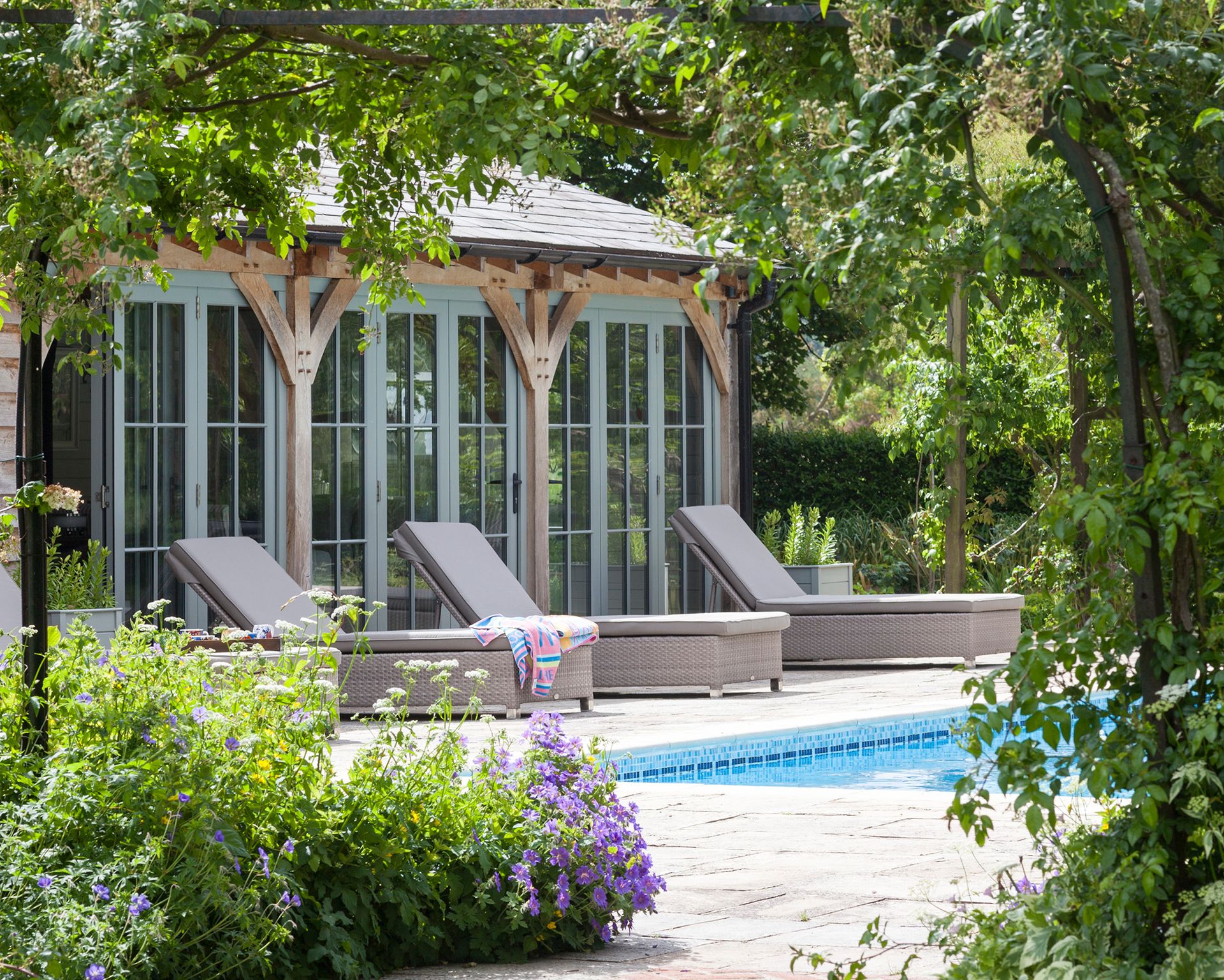 A pool area with sun loungers next to a large pool house with french doors