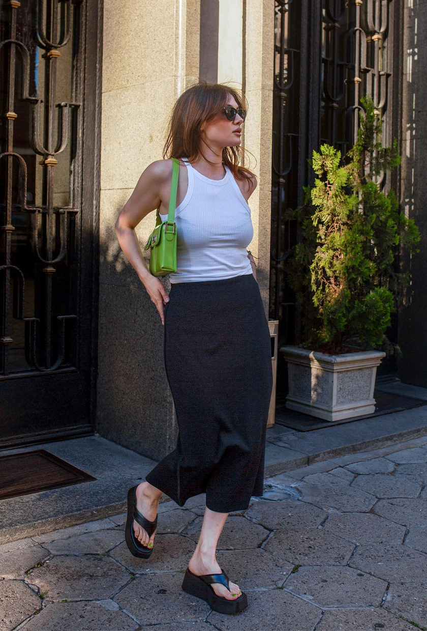 The sandal trend wedge flip-flops are shown in a photo of a woman standing outside wearing a white tank top, a black slip shirt, black wedge flip-flop heels, a green shoulder bag, and black sunglasses