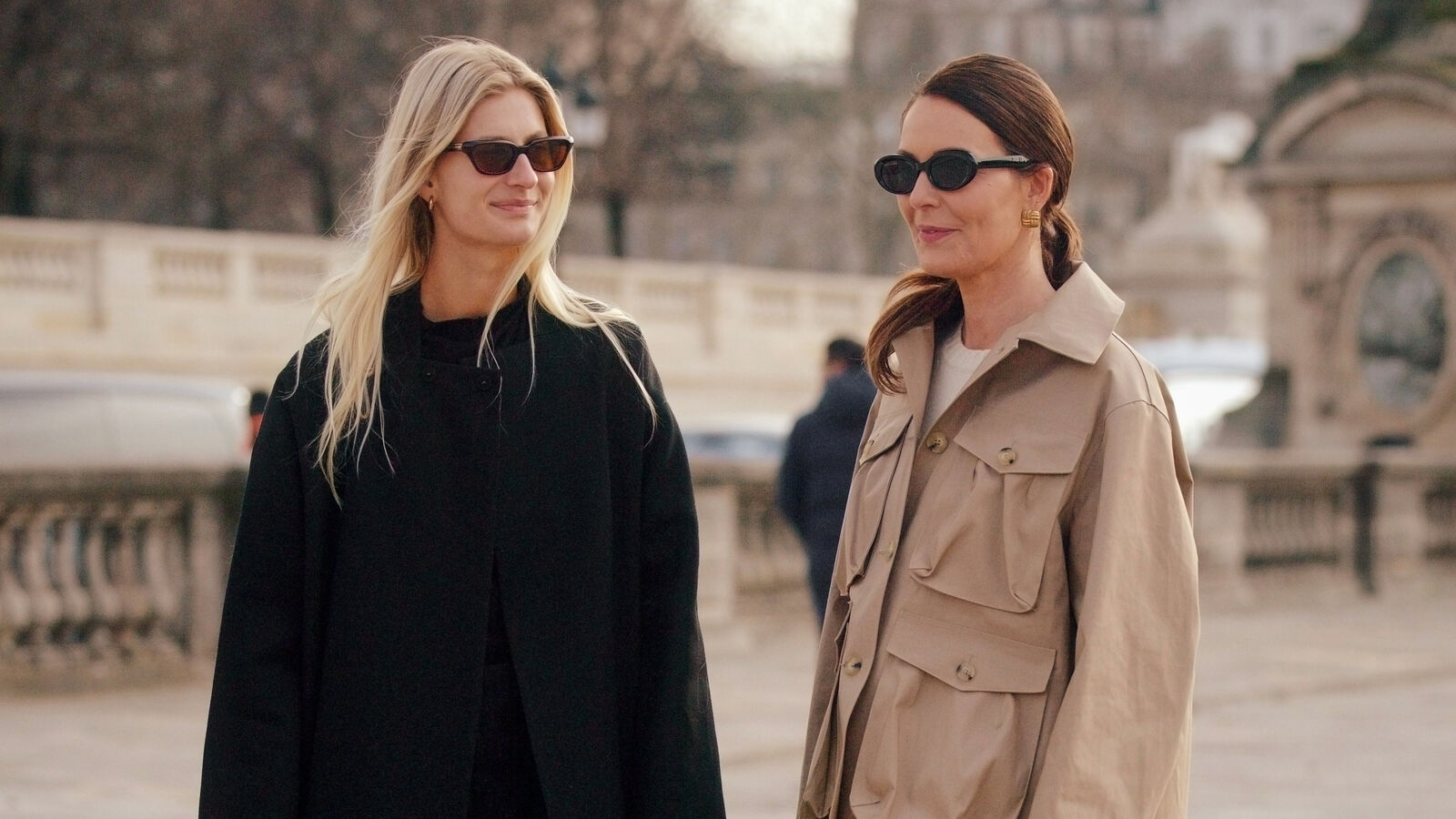 Two women pose while speaking to each other in Paris. The one on the left is wearing an all black outfit with sunglasses and an Issey Miyake handbag. The woman on the left is wearing a khaki jacket and black pants with a white handbag and sunglasses. 