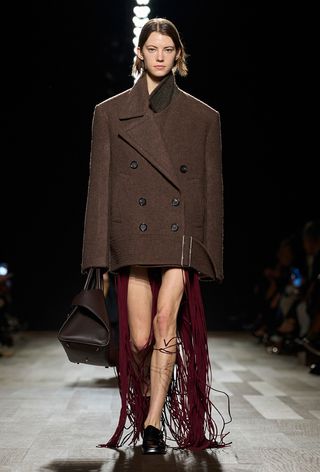 A model from Ferragamo's fall 2024 runway wearing a brown peacoat over a red fringe dress styled with a brown bag and brown loafers.