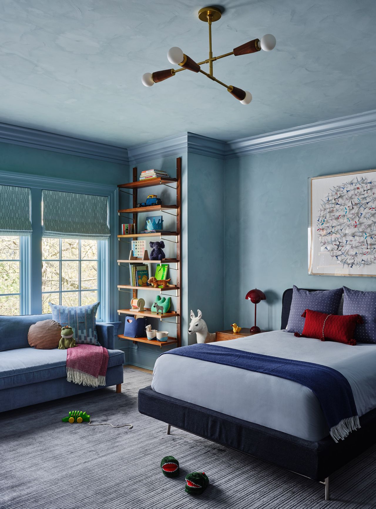 A light blue color drenched modern kids' room with a large bed, sofa, and bookshelves with a piece of art hanging behind the bed, and some toys on the floor and on the sofa with a statement ceiling light