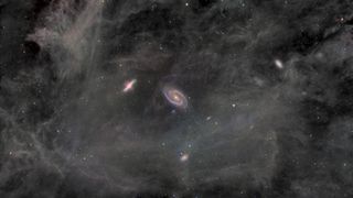 The spiral galaxy M81 is pictured to the right of the Cigar Galaxy against the cloud-like backdrop of a nebula. Other galaxies and stars punctuate the image. The spiral galaxy M81 features sweeping spiral arms and a bright core.