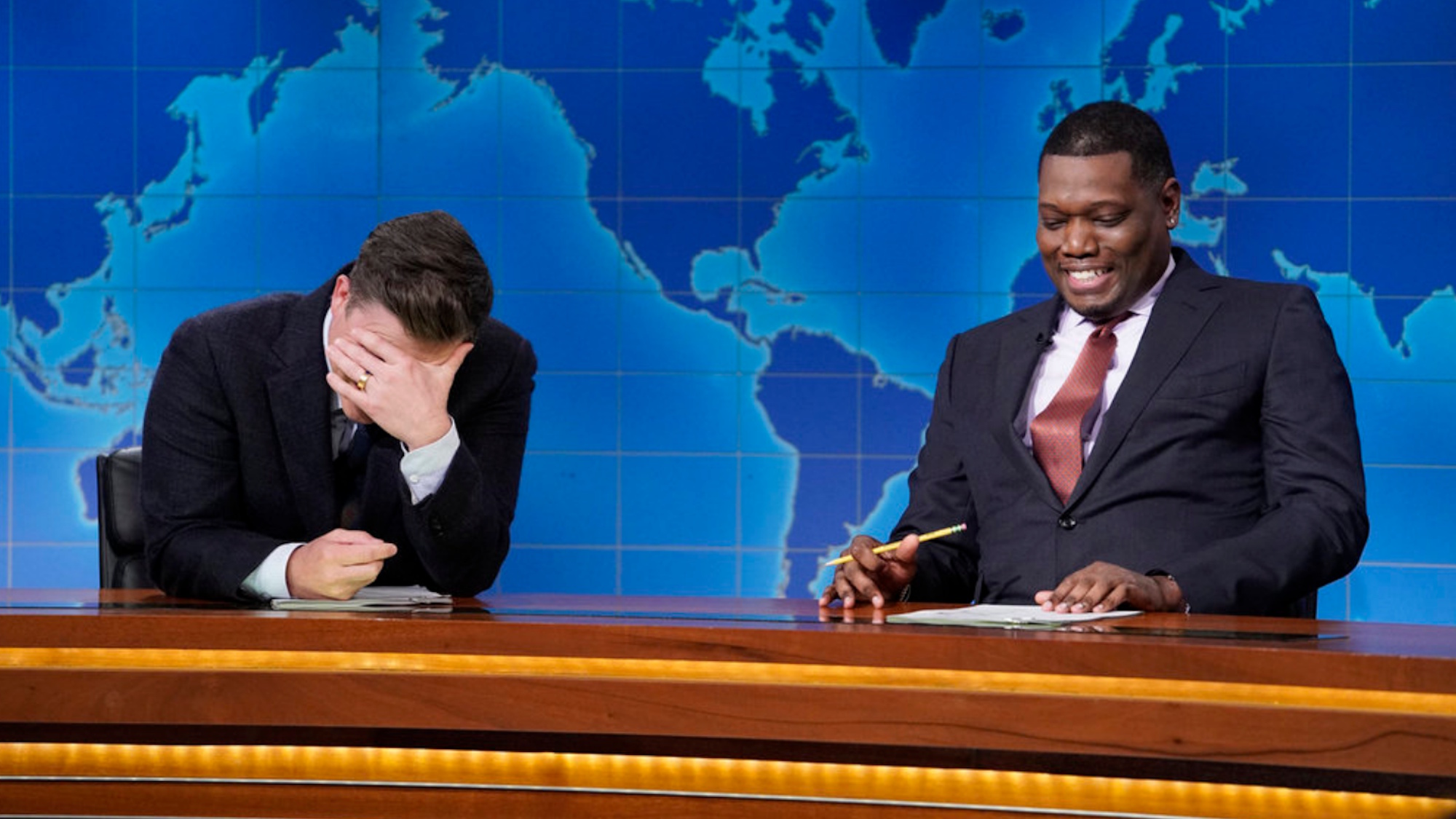 Colin Jost hides his face in his hands as Michael Che laughs during one of their joke swaps on Saturday Night Live's Weekend Update.