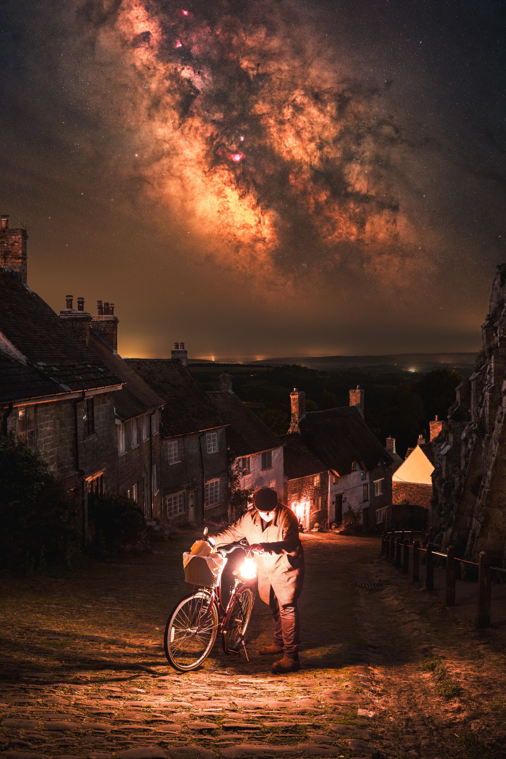 A man is photographed pushing a bike up a cobbled hill lined by houses as the Milky Way glows brightly overhead at night.