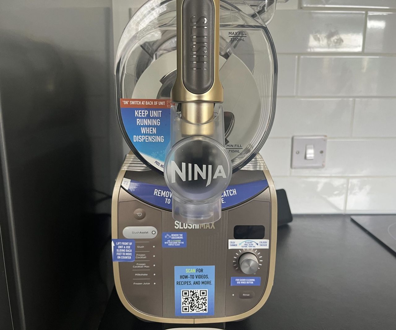 The front of the gold and grey Ninja Slushi Max with the blue instruction stickers on it, on a black kitchen countertop.
