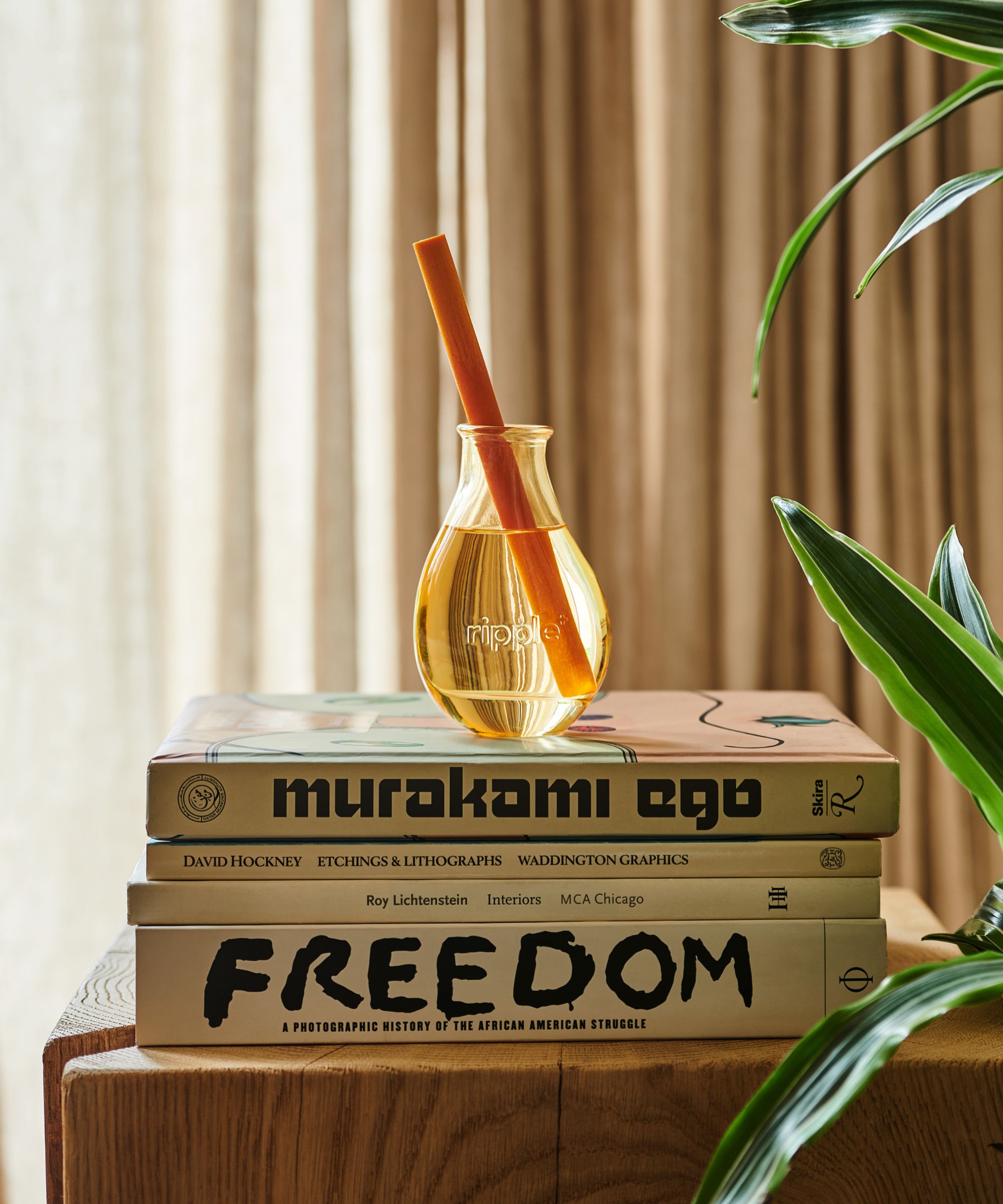 A ripple+ diffuser on a stack of books on top of a wooden surface with neutral curtains behind and leaves to the right