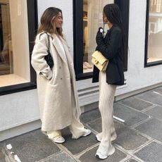 European influencer Amaka Hamelijnck and Bruna Rizk pose on a sidewalk in Paris wearing chic winter outfits with a long cream textured coat, a blazer with ribbed pants, and sneakers