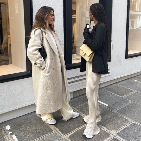 European influencer Amaka Hamelijnck and Bruna Rizk pose on a sidewalk in Paris wearing chic winter outfits with a long cream textured coat, a blazer with ribbed pants, and sneakers