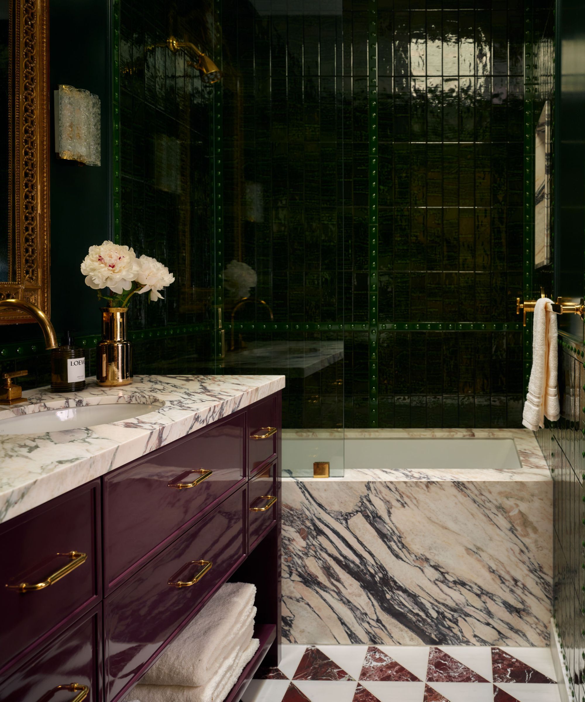 a glossy dark green tiled bathroom with a lacquered purple vanity, marble bath and floor tiles with brass accessories and hardware