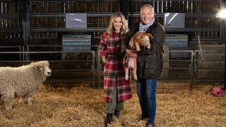 Helen Skelton and Jules Hudson in a barn with Jules holding a piglet and a sheep looking on in Winter on the Farm.