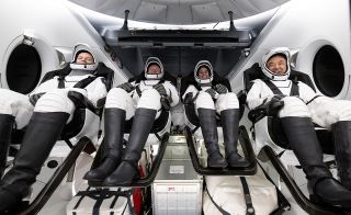 four people in black and white spacesuits sit inside a white space capsule