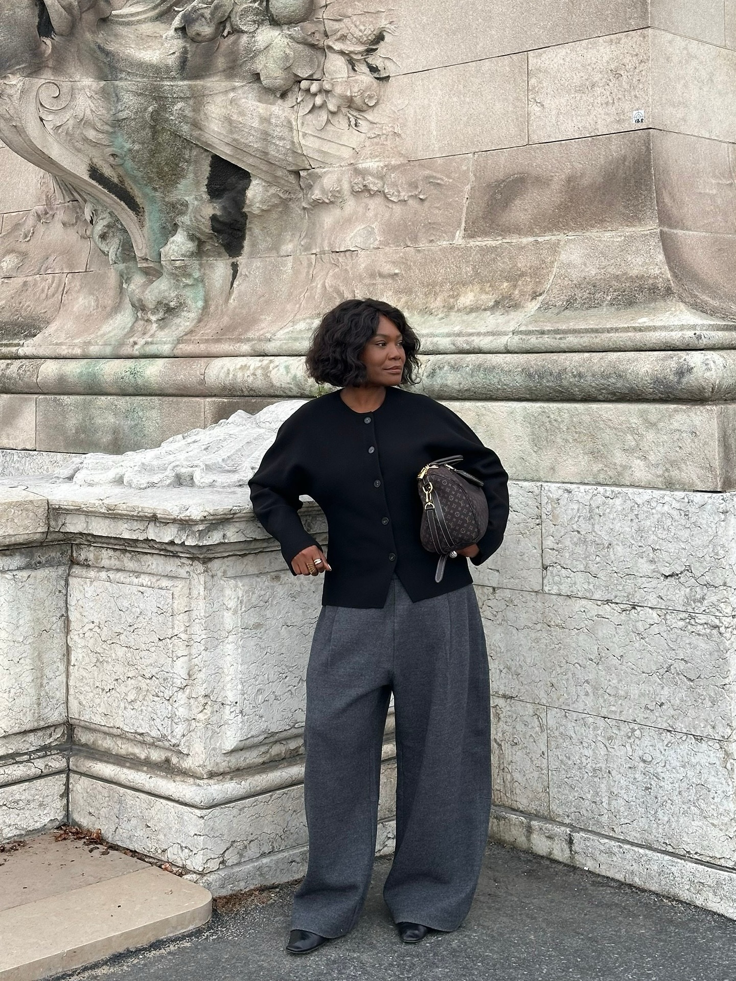 Franny wears a black cardigan, grey tailored trousers, black boots