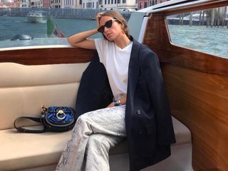 Italian fashion influencer Erika Boldrin poses while sitting on a boat in Venice wearing black cat-eye sunglasses, red earrings, a white t-shirt, oversize black blazer, black patent bag, and snakeskin pants