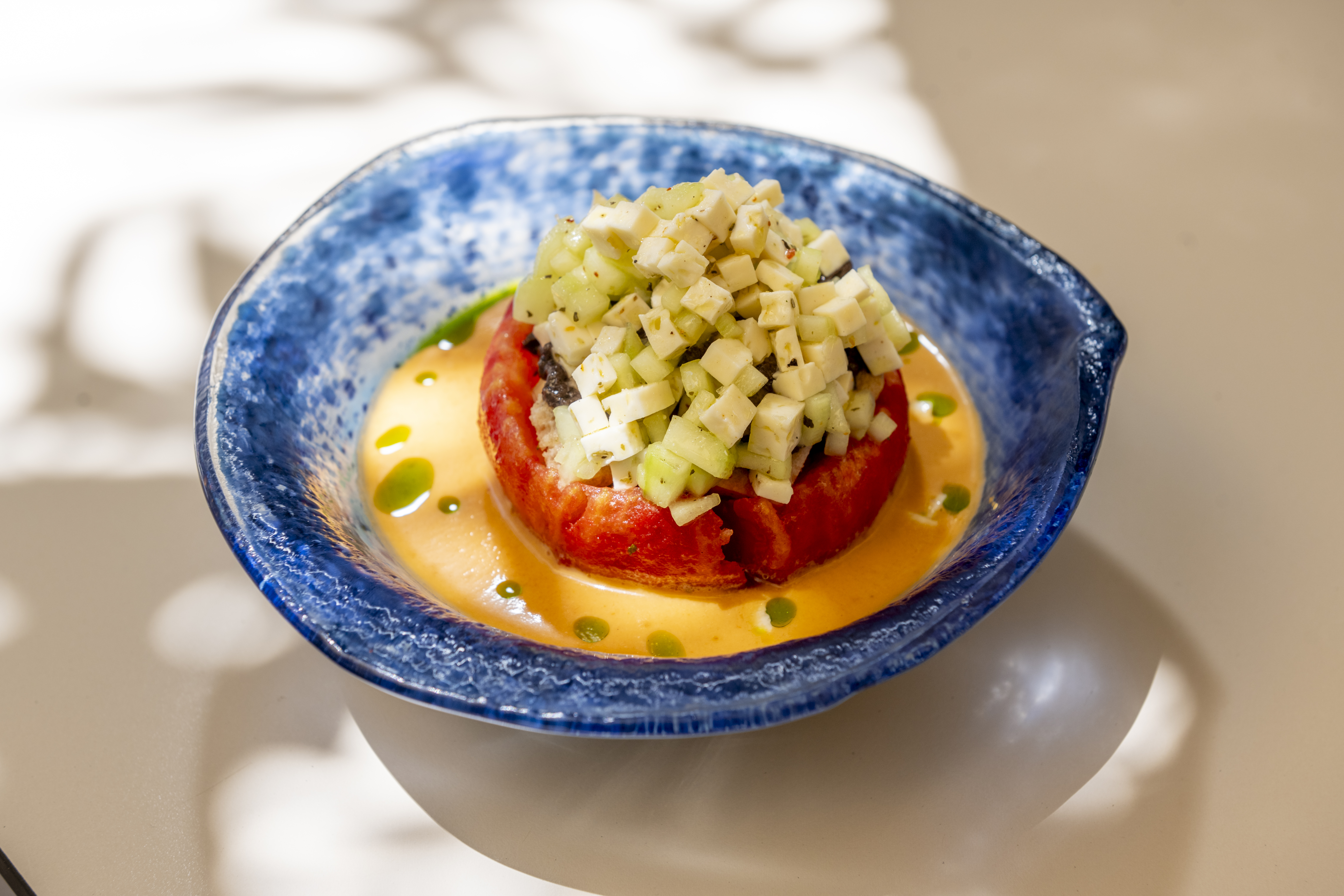A glazed, organically shaped blue bowl sits in the sunshine as a tartare rests on a layer of yellow sauce, topped with diced vegetables and cheese.