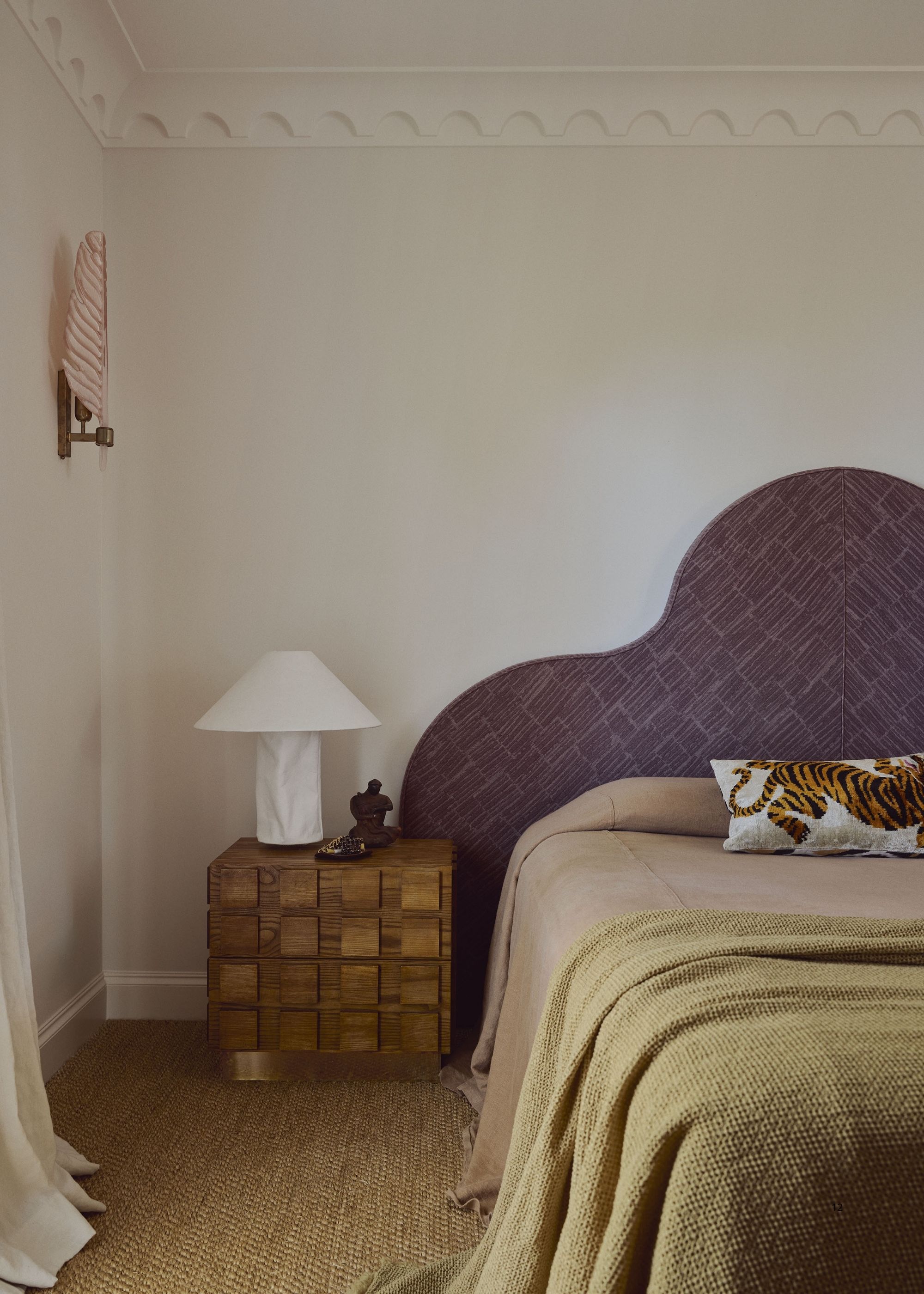 An aubergine upholstered headboard in a white bedroom with a tan bedspread and a green throw. 