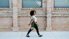 A woman in a t-shirt, leggings and sandals walks outside. She is smiling and swinging her arms gently. Behind her we see a brick building. 