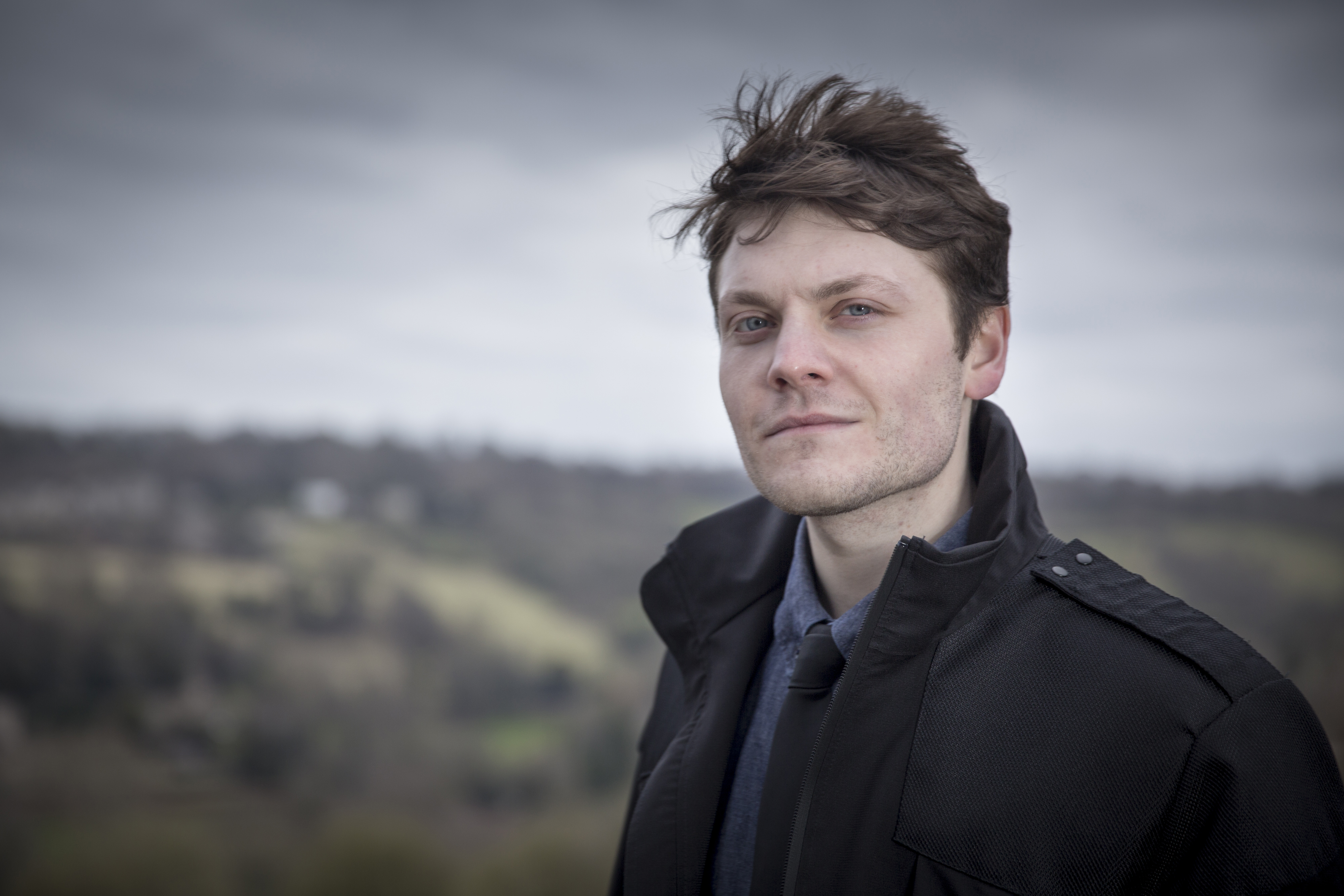 White man with dark hair, shirt and tie and black jacket, posing against a backdrop of a hillside and sky