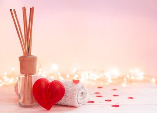 A diffuser and white flannel rolled up, with a red heart shaped soap and red heart confetti in front of a light pink background and fairy lights.