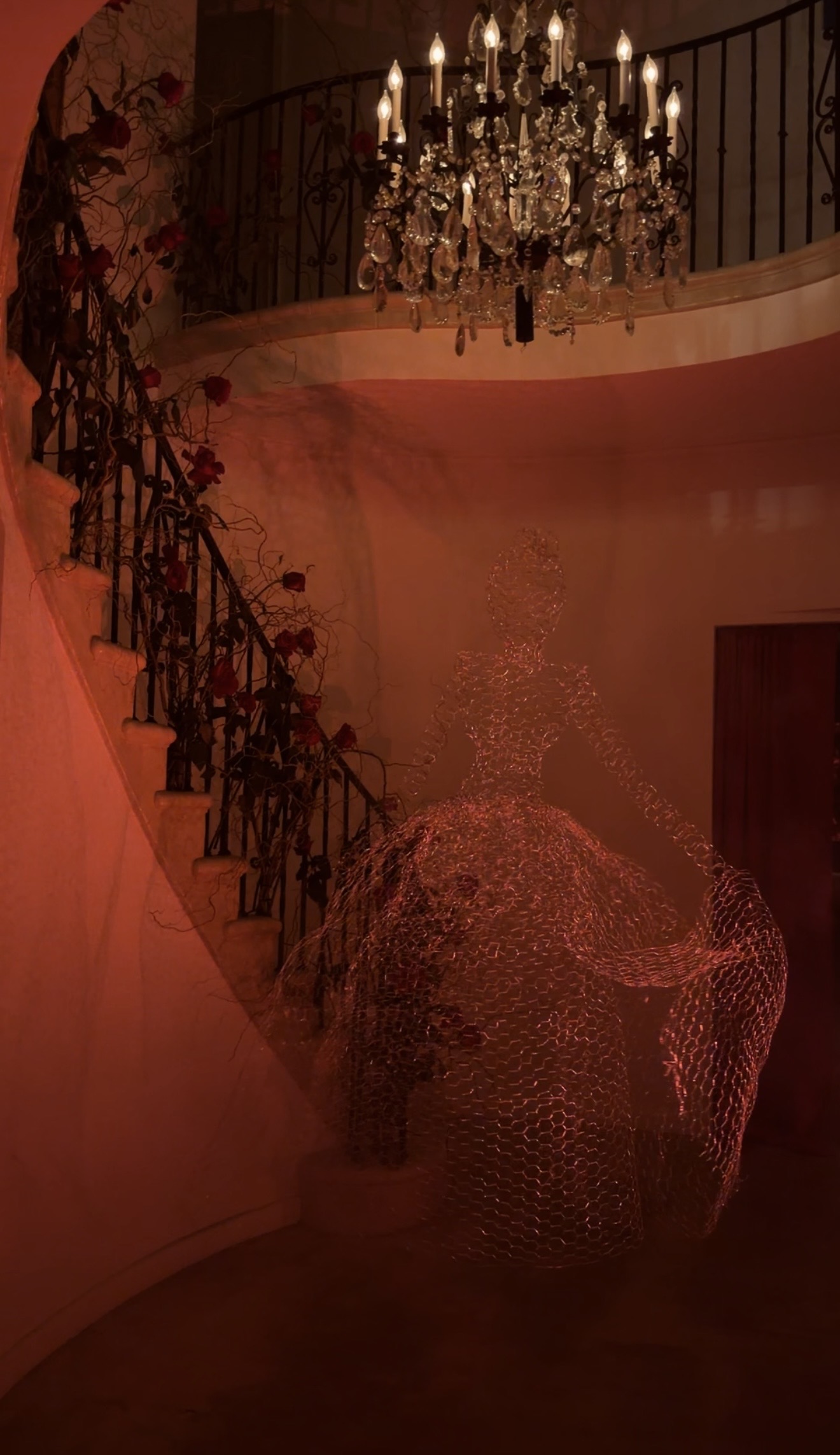 An entryway decorated for Halloween lit with a red light and with a floating ghost made of chicken wire under the chandelier at the foot of a spiral staircase adorned with red roses and branches