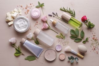 A variety of beauty products with flowers and green stems on a light pink background.