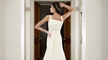 Girl wearing a white wedding dress with a bow, photographed from behind with one arm of her hip and the other on her head