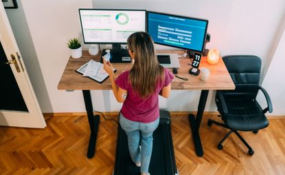 A woman walks on a treadmill while working at her desk. She wears jeans, socks and a t-shirt. There are two screens on her desk along with a plant and some papers. A wheeled chair sits to the side of the desk.