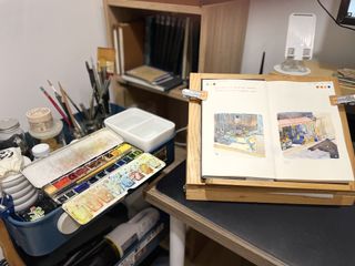 Artist sketchbook; an open sketchbook on a drawing table, in an artist's studio, to the side is a watercolour paints palette