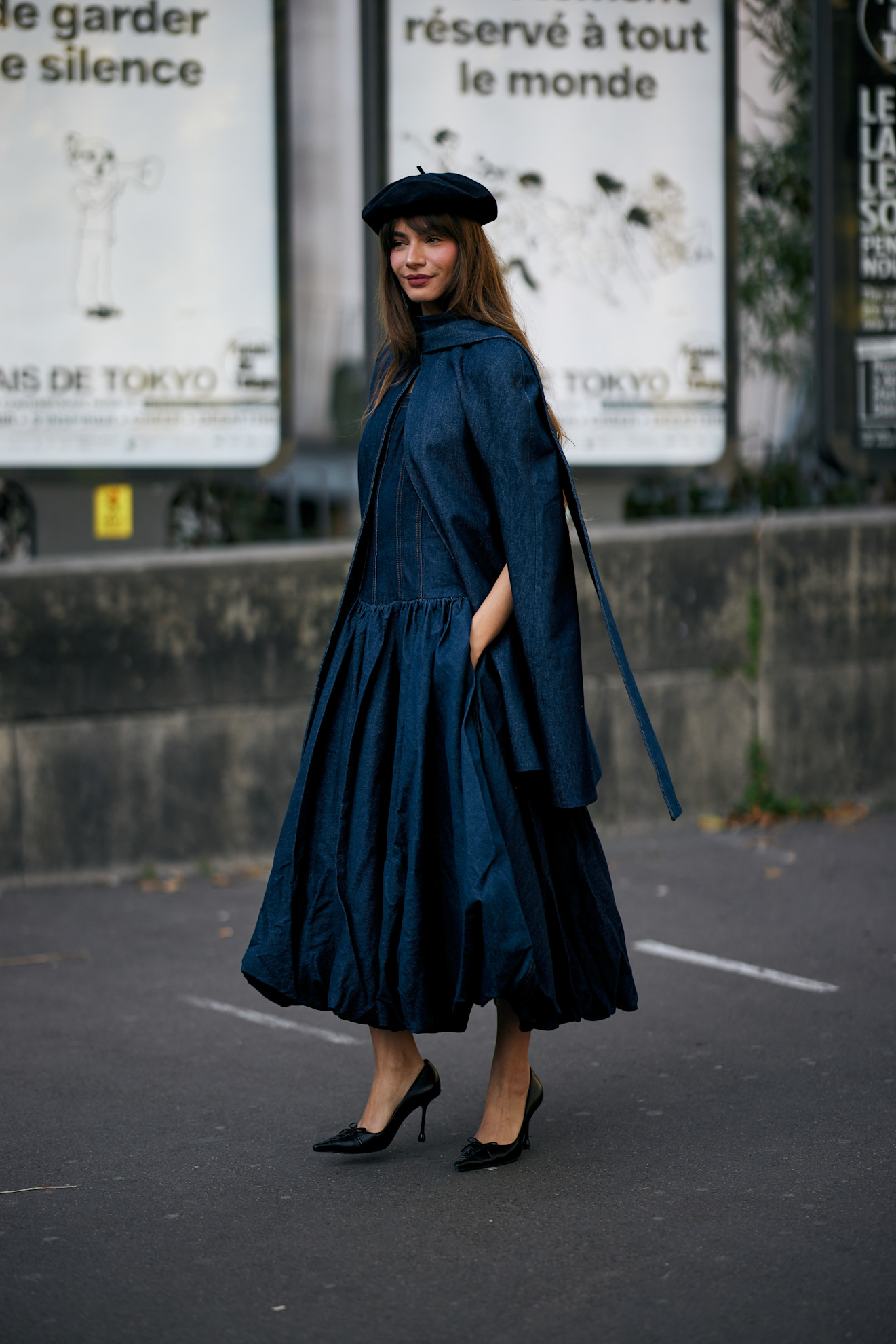 Woman in a denim bubble skirt dress, denim cape, black beret, and black stilettos.