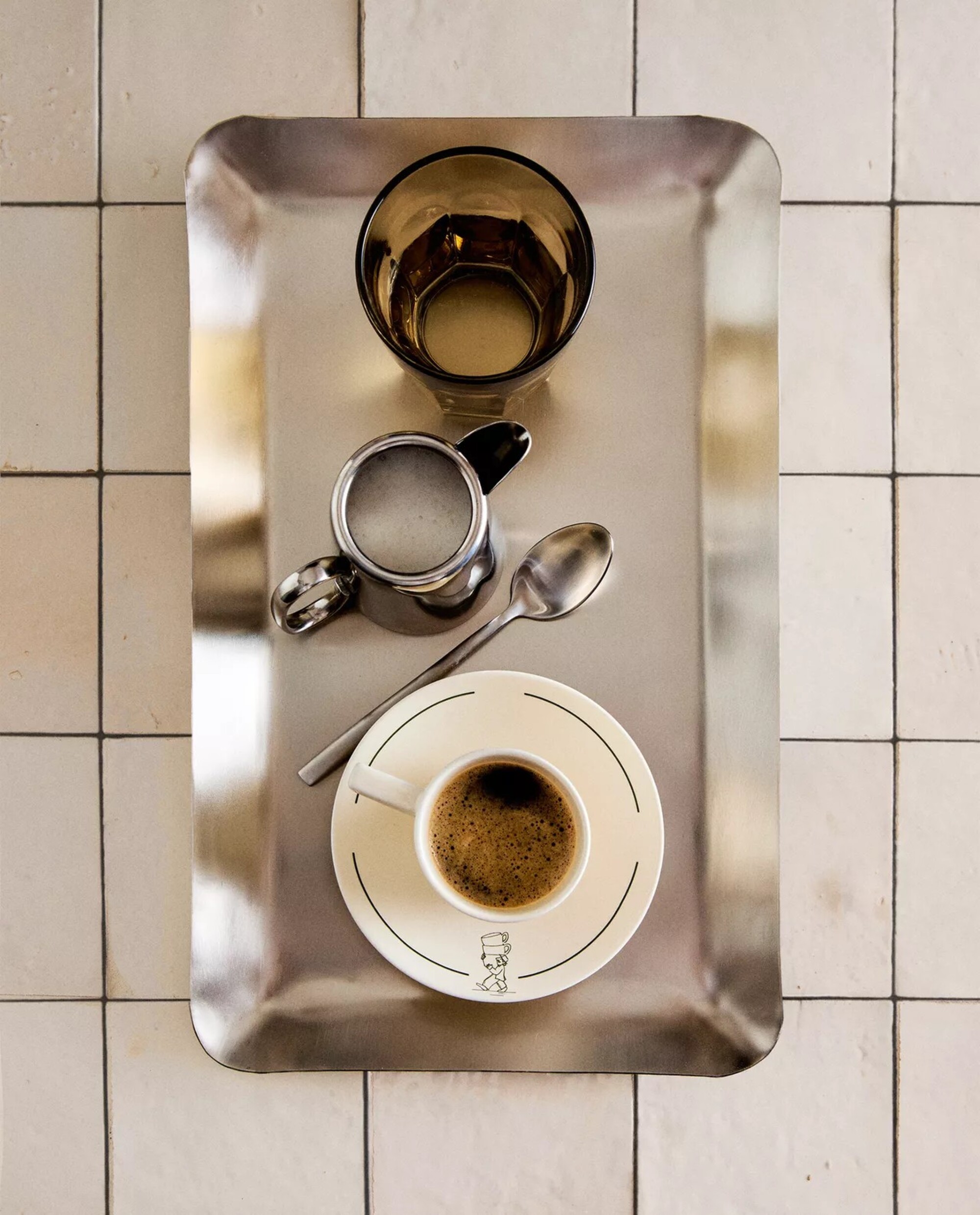 A silver tray on a white tile countertop. A white espresso cup and saucer, a glass cup, a silver milk cup and spoon are on the tray.