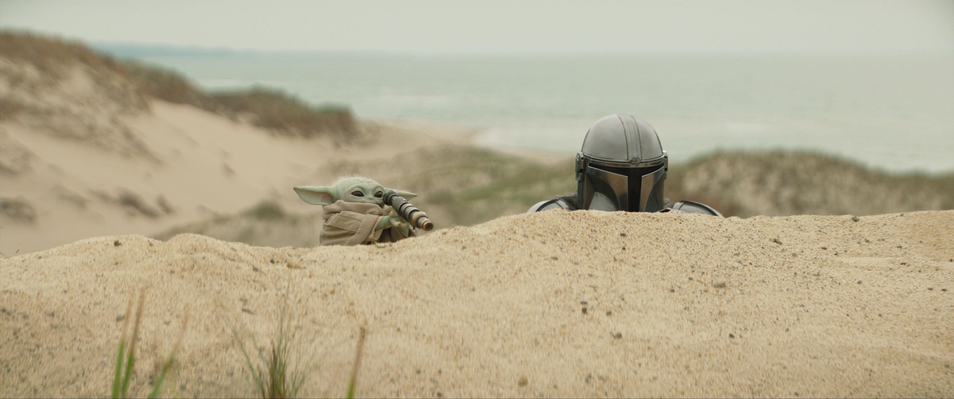 Screenshot from The Mandalorian &amp; Grogu, showing the titular characters hiding behind a sand dune.