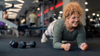 A smiling woman in sportswear performs a plank in a gym. Her body is elevated off the floor, held in a straight line from her shoulders to her heels, and her weight is supported by her forearms and tiptoes. In the background we see gym equipment and other gym goers.