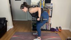 Freelance fitness writer Jennifer Rizzuto performs a dumbbell workout. She is in a living room, with a book case and kettlebells behind her and a yoga mat on the floor. She is hinging over at her hips so her torso is nearly parallel with the floor, a pair of dumbbells grasped in each hand, with her elbows bent and hands held close to her hips. 