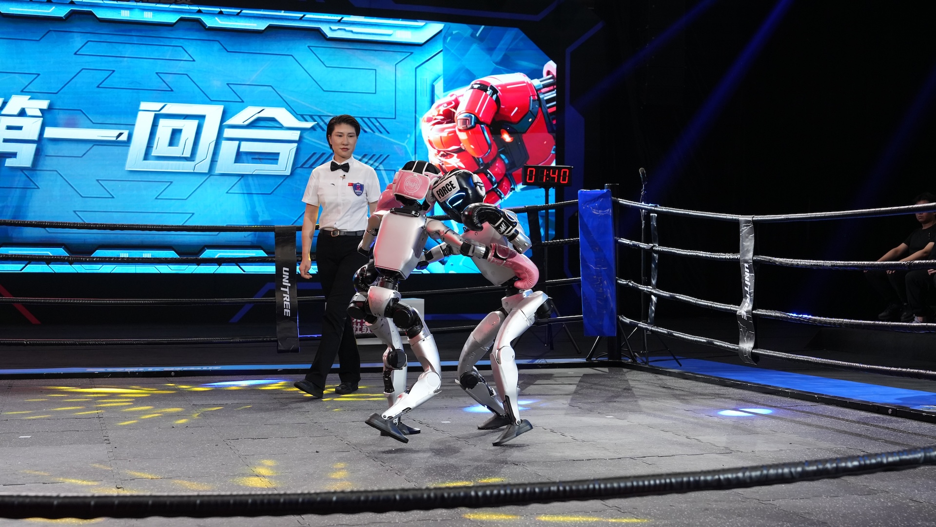 Two robots fight in a boxing match as an announcer watches
