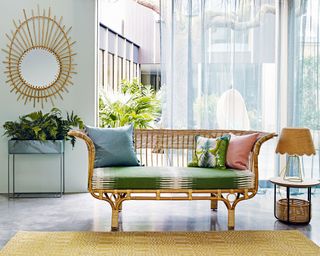 Belladonna rattan sofa with green seat cushion on screed floor in front of window with light blue sheer curtains, basket side table and rattan mirror on wall.