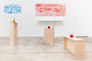 In a brightly lit modern gallery, prints in the tones of electric blue and red hang on otherwise white walls while, in front of them, apples and pears sit on wooden podiums and benches.