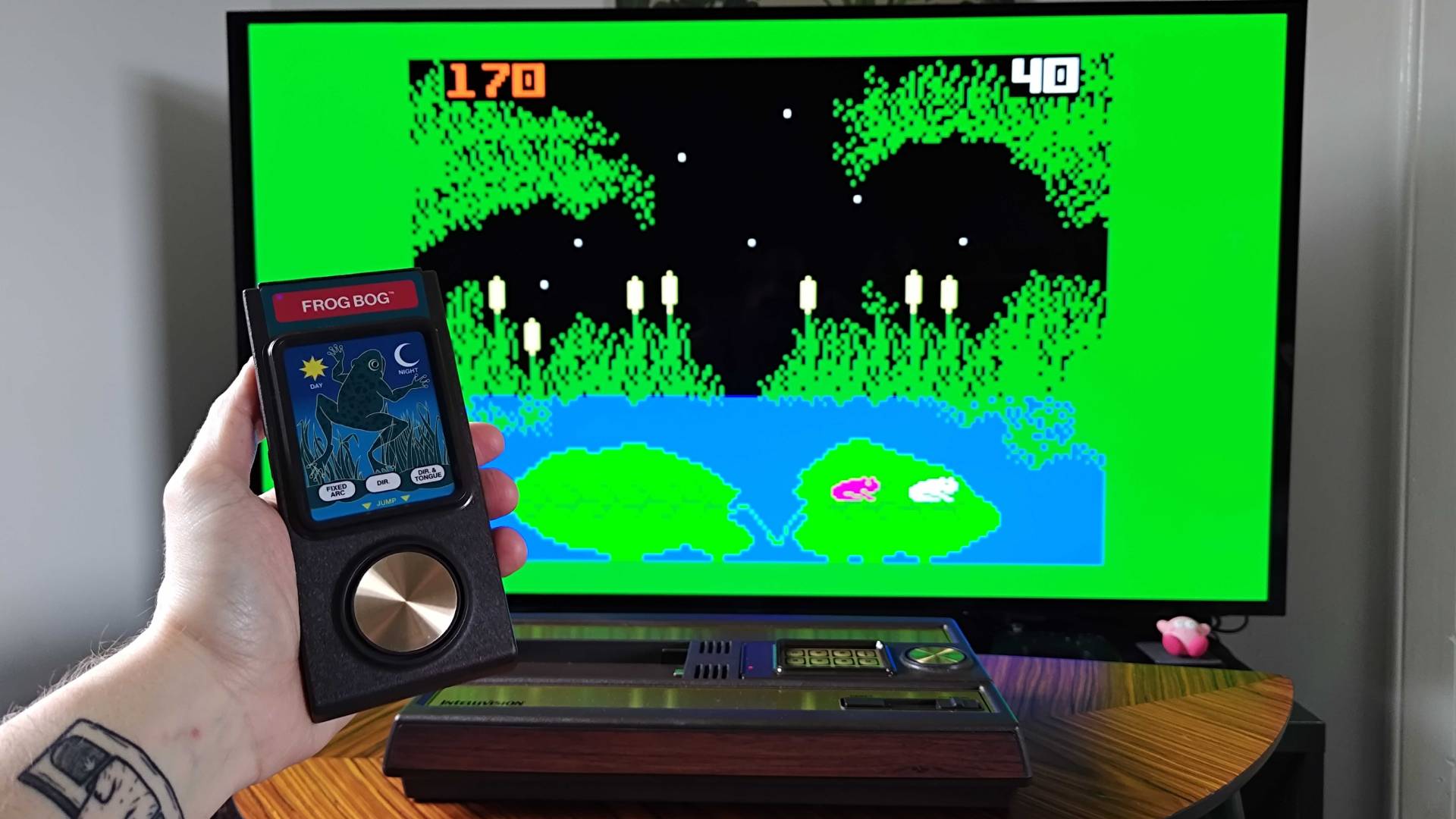 Hand holding Intellivision Sprint controller with Frog Bog gameplay in backdrop.