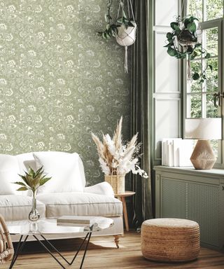 A green living area with green floral wallpaper, hanging plants, a tall window with a ledge with a rattan lamp, a white couch with pampas grass next to it and a coffee table in front of it with dried flowers and light wooden flooring