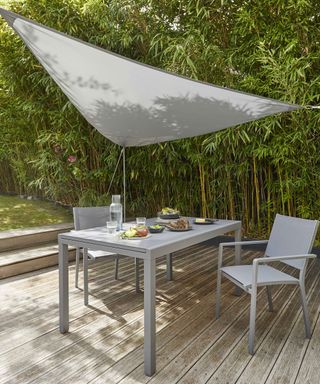 A grey 3x3m shade sail above a grey garden dining set on a decked area surrounded by bamboo plants