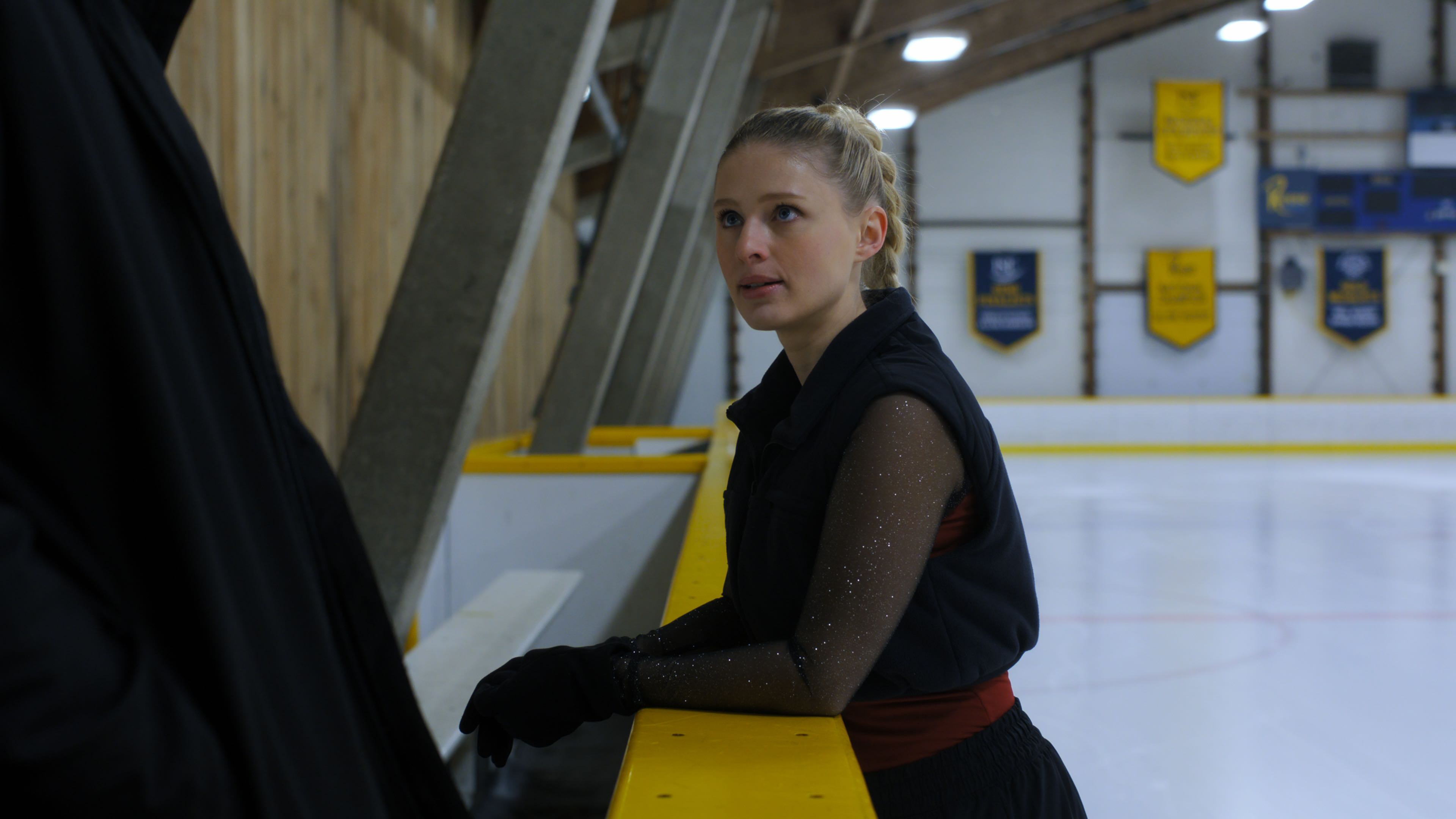 Alexandra Beaton as Elise Russo wearing a vest and long sleeves while standing on the ice and leaning over the side of the rink in Finding Her Edge season 1
