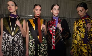 Four female models wearing looks from Prada's collection. One model is wearing a green, black, gold and patterned piece and purple neck scarf. The model next to her is wearing a light orange, black and patterned piece and brown neck scarf. The third model is wearing a light purple neck scarf, red, black, grey and white pussybow blouse and a black and brown striped piece over the top. And the fourth model is wearing a yellow, brown and black pussybow blouse and purple neck scarf. All four models are holding bags with chain handles on their shoulders