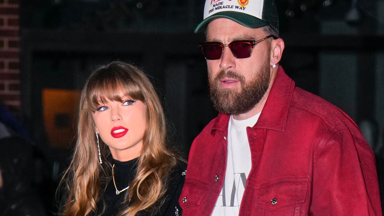 Taylor Swift has bright red lipstick and brown hair as she walks with Travis Kelce who is wearing a baseball cap