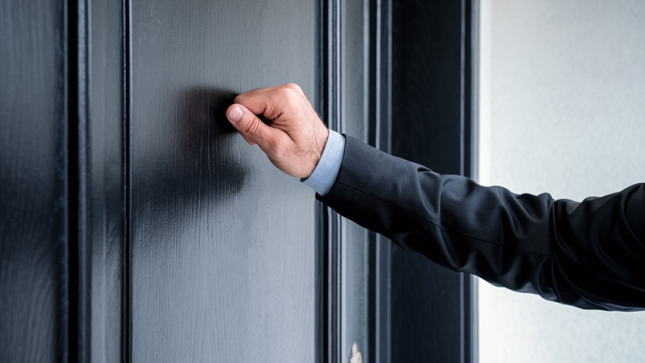 We see the arm of a man wearing a suit knocking on a blue door.