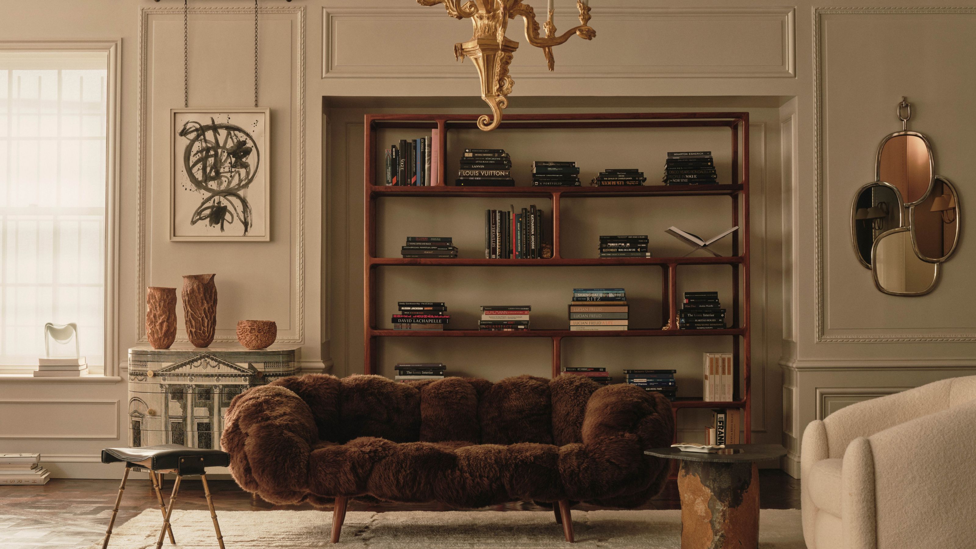 Cream living room with paneled walls and modern furniture, including a brown furry couch, a wooden minimalist bookcase and a cream bouclé arm chair