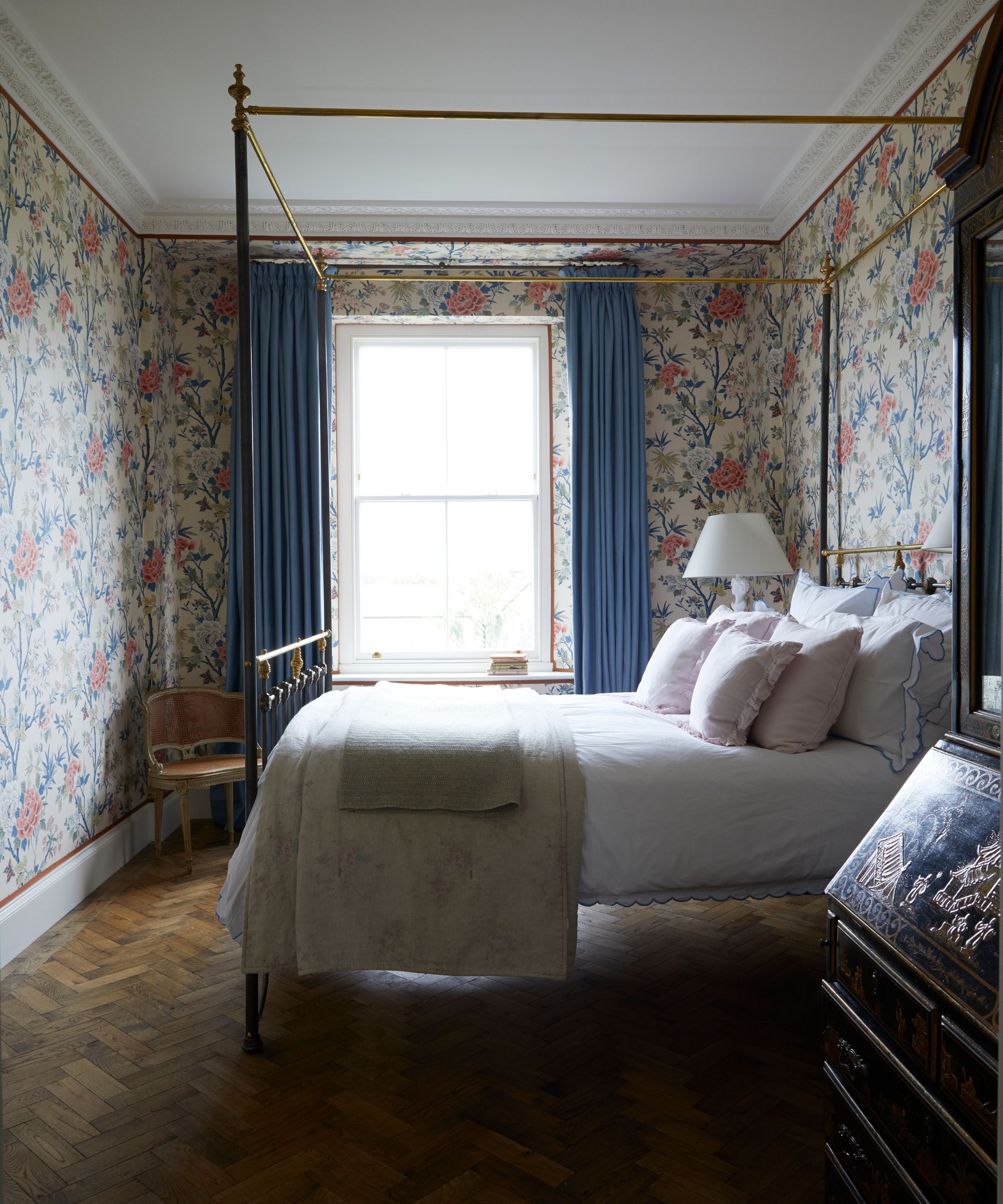 A floral bedroom with a large four poster bed with white bedding, beige blankets, and pale pink cushions. In the background is a large window with open blue curtains, and in the foreground is a dark wood engraved dresser. In the far left corner is a rattan chair.