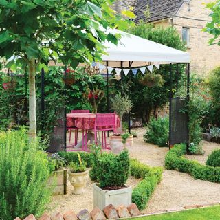 Black gazebo with white fabric covering above a pink outdoor dining set on a gravel area zoned with low level shrubs