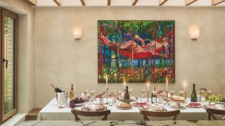 A large rectangular diing table covered in a white tablecloth with lit taper candles, bottles of champagne and champagne glasses and table settings ready for dining under a large colorful artwork