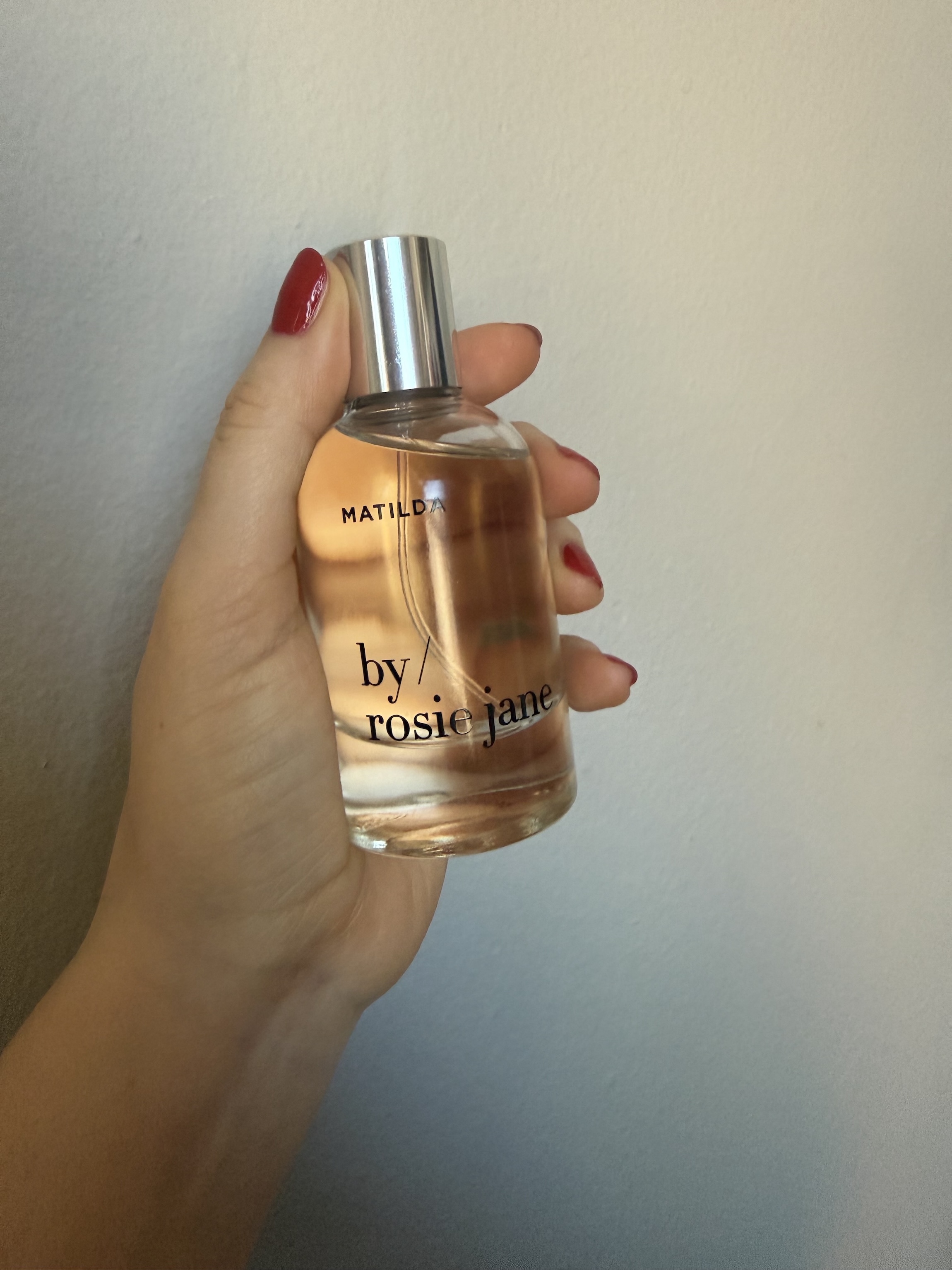 hand holding up by rosie jane matilda perfume bottle against grey wall